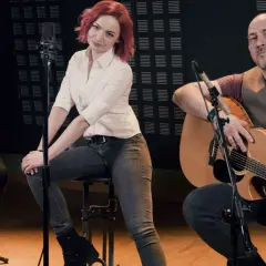Zespół wykonuje brathanki utwory. Dwóch mężczyzn gra na gitarach, a kobieta z rudymi włosami siedzi pośrodku.