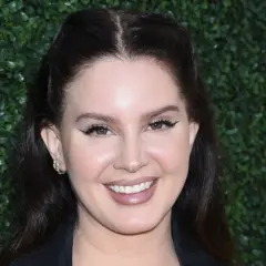 Uśmiechnięta wokalistka Lana Del Rey z charakterystycznym makijażem oczu i subtelną biżuterią na uchu.