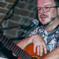 Jacek Kaczmarski z gitarą i mikrofonem, w okularach i wzorzystej koszulce. Prywatne chwile artysty.
