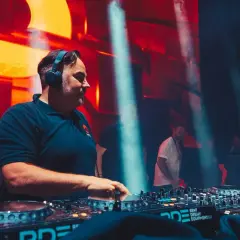 DJ Kris w akcji, miksuje muzykę na żywo. W tle pulsujące światła i tłum.