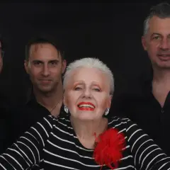 Edek Tango i jego zespół w czerniach, gotowi na występ. Wokalistka z czerwoną broszką błyszczy na pierwszym planie.