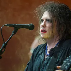 Robert Smith, the iconic wokalista The Cure, śpiewa do mikrofonu, z charakterystycznym makijażem i rozczochranymi włosami.