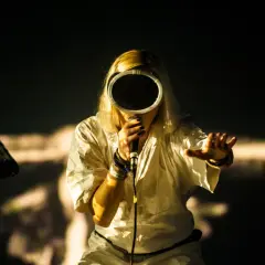 Artystka z maską na twarzy, trzymająca mikrofon. Na scenie widać sprzęt muzyczny, a w tle światła. To jej bokka skład.