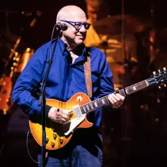 Mark Knopfler, w okularach i niebieskiej koszuli, gra na gitarze Gibson. Jego muzyka, jak na jego albumy, brzmi w tej chwili na żywo.