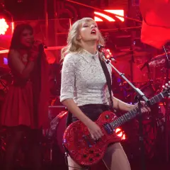 Taylor Swift gra na czerwonej gitarze podczas koncertu. W tle widać perkusję i inną wokalistkę. To może być fragment trasy promującej jej taylor swift albumy.