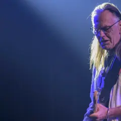 Muzyk z zespołu Lipińskiego w okularach i z długimi włosami, grający na gitarze podczas koncertu.