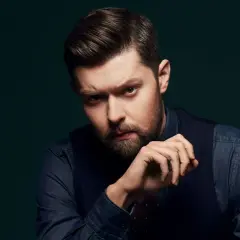 Mężczyzna z brodą i starannie ułożonymi włosami, w ciemnej koszuli i kamizelce, spogląda intensywnie. Jego dłoń opiera się o podbródek, jakby zastanawiał się nad kolejnymi mrozu utworami.
