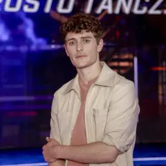 Młody mężczyzna z kręconymi włosami, w beżowej koszuli, stoi na tle sceny. Jakub Józef Orliński wzrostem imponuje, ale jego talent jest jeszcze większy.