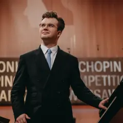 Młody pianista, jeden z polskich laureatów Konkursu Chopinowskiego, w eleganckim garniturze przy fortepianie.