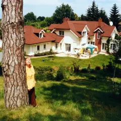 Zdzisława Sośnicka, uśmiechnięta, opiera się o drzewo na tle swojego domu. Jej pochodzenie jest polskie.