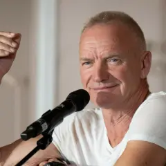 Sting, wokalista zespołu The Police, przemawia do mikrofonu, unosząc rękę w geście.