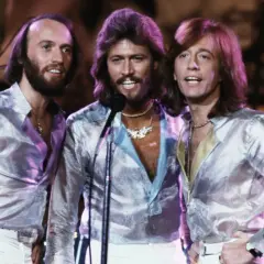 Bracia Bee Gees w błyszczących koszulach i białych spodniach, śpiewający na scenie.