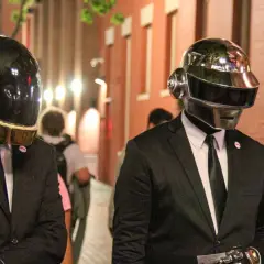 Daft Punk członkowie w złotych i srebrnych hełmach, ubrani w garnitury, idą ulicą.