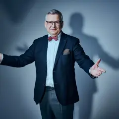 Artur Andruś, w eleganckim garniturze i muszce, gestykuluje, jakby opowiadał o swojej rodzinie.