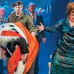Król opera Pendereckiego w pomarańczowym płaszczu z futrem kłania się nisko. Obok kobieta w błyszczącej sukni.