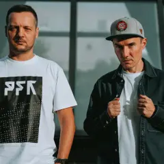 Dwóch członków Paktofoniki, jeden w białej koszulce z logo, drugi w czarnej kurtce i czapce z daszkiem.
