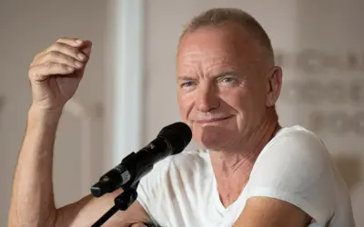Sting, wokalista zespołu The Police, przemawia do mikrofonu, unosząc rękę w geście.