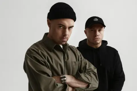 Dwóch mężczyzn, jeden w czarnej czapce beanie i oliwkowej koszuli, drugi w czarnej czapce z daszkiem. Zapowiedź nowych pro8l3m albumy.