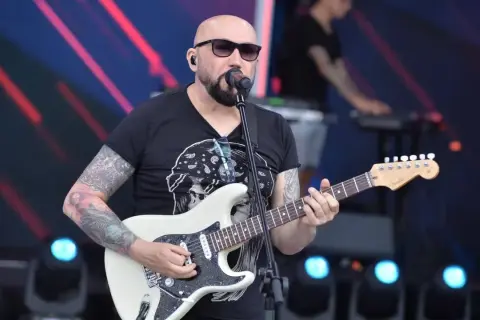 Artysta z tatuażami gra na gitarze, śpiewając do mikrofonu. Koncertowy klimat, jakby to były **kombi piosenki alfabetycznie**.