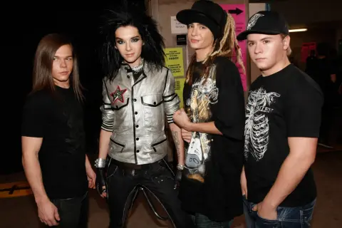 Członkowie Tokio Hotel pozują do zdjęcia. Bill Kaulitz w błyszczącej kurtce, Tom Kaulitz w czapce i z dredami, a obok nich Georg Listing i Gustav Schäfer.