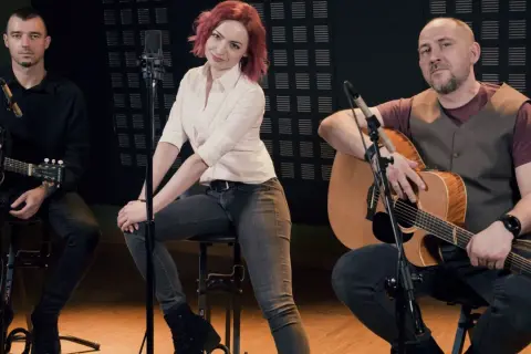 Zespół wykonuje brathanki utwory. Dwóch mężczyzn gra na gitarach, a kobieta z rudymi włosami siedzi pośrodku.