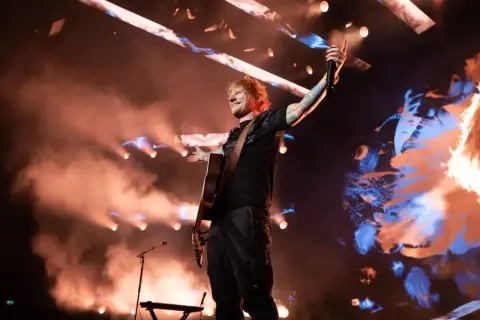 Ed Sheeran w Polsce, na scenie z gitarą, w otoczeniu dymu i świateł, pozdrawia publiczność.