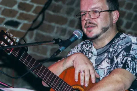 Jacek Kaczmarski z gitarą i mikrofonem, w okularach i wzorzystej koszulce. Prywatne chwile artysty.