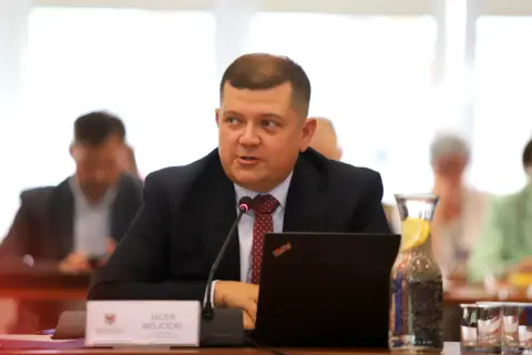Jacek Wójcicki, siedząc przy stole, przemawia. Jego wzrost jest przeciętny, a na stole leży laptop i woda z cytryną.