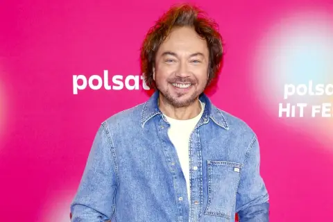 Mietek Szczęśniak, uśmiechnięty, w dżinsowej koszuli, na tle różowego banera z logo Polsat Hit Festiwal.