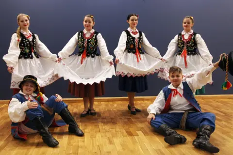 Dzieci w strojach ludowych, dziewczęta trzymają białe spódnice, chłopcy w niebieskich spodniach i kamizelkach, gotowi do tańca kołomyjka.