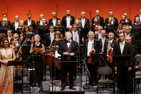 Orkiestra i chór przygotowują się do wykonania **Moniuszko opery**. Dyrygent stoi na podwyższeniu, gotowy do rozpoczęcia.
