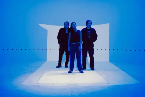 Trzy osoby stoją na scenie oświetlonej na niebiesko, tworząc atmosferę idealną do shoegaze.