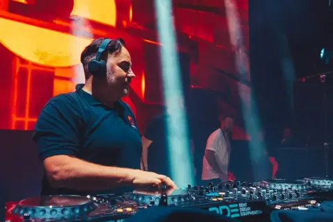 DJ Kris w akcji, miksuje muzykę na żywo. W tle pulsujące światła i tłum.