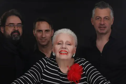 Edek Tango i jego zespół w czerniach, gotowi na występ. Wokalistka z czerwoną broszką błyszczy na pierwszym planie.