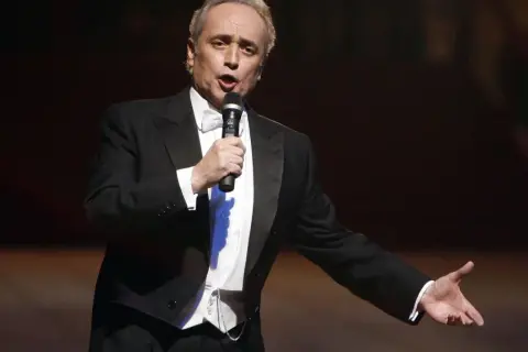 José Carreras, hiszpański tenor, w eleganckim smokingu śpiewa do mikrofonu, gestykulując.