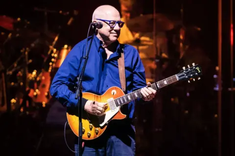 Mark Knopfler, w okularach i niebieskiej koszuli, gra na gitarze Gibson. Jego muzyka, jak na jego albumy, brzmi w tej chwili na żywo.