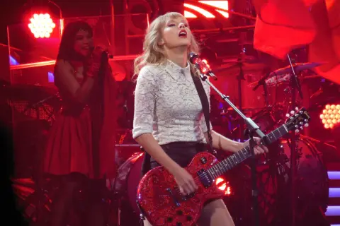 Taylor Swift gra na czerwonej gitarze podczas koncertu. W tle widać perkusję i inną wokalistkę. To może być fragment trasy promującej jej taylor swift albumy.