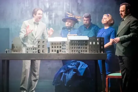 Aktorzy na scenie, prezentujący polskie musicale, gestykulują przy makiecie miasta.