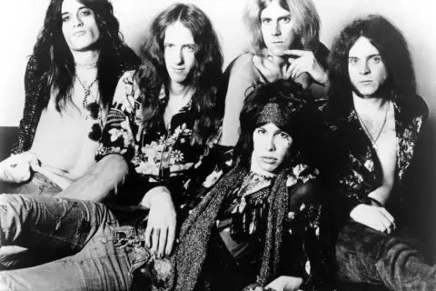 Aerosmith członkowie w rockowym stylu, z długimi włosami i charakterystycznymi strojami.