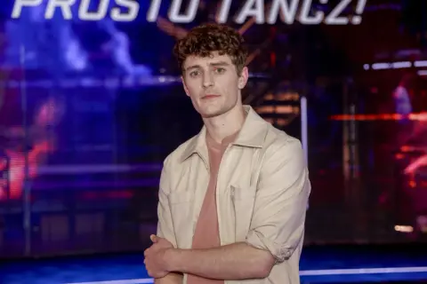 Młody mężczyzna z kręconymi włosami, w beżowej koszuli, stoi na tle sceny. Jakub Józef Orliński wzrostem imponuje, ale jego talent jest jeszcze większy.