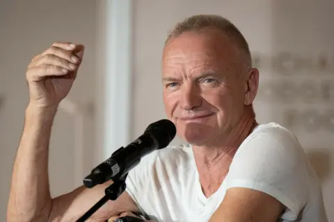 Sting, wokalista zespołu The Police, przemawia do mikrofonu, unosząc rękę w geście.