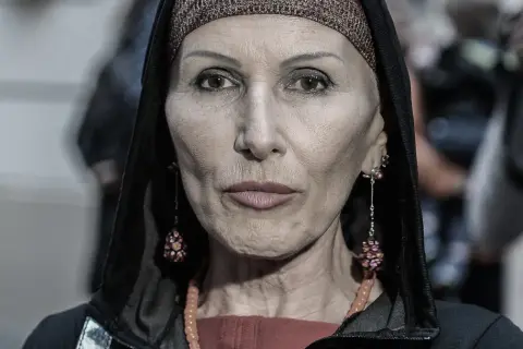 Kobieta w kapturze i ozdobnej opasce na głowie. Jej twarz, naznaczona kora wiek, opowiada historię. Długie kolczyki i naszyjnik dodają jej charakteru.