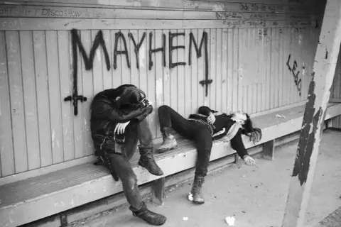 Dwie postacie w mrocznym stylu, leżące na ławce pod napisem "MAYHEM". Może to być okładka jednego z albumów zespołu Mayhem.