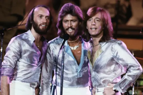 Bracia Bee Gees w błyszczących koszulach i białych spodniach, śpiewający na scenie.