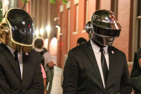 Daft Punk członkowie w złotych i srebrnych hełmach, ubrani w garnitury, idą ulicą.