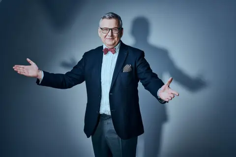 Artur Andruś, w eleganckim garniturze i muszce, gestykuluje, jakby opowiadał o swojej rodzinie.