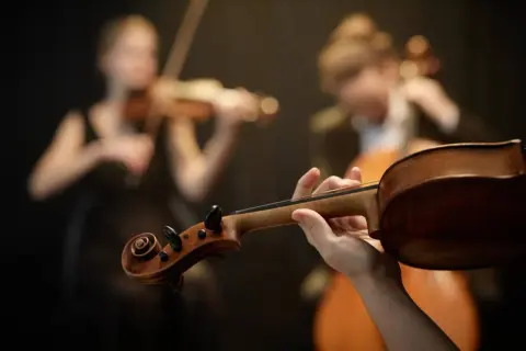 Dłoń trzymająca gryf skrzypiec, w tle dwie kobiety grające na instrumentach smyczkowych. To preludium do pięknej muzyki.