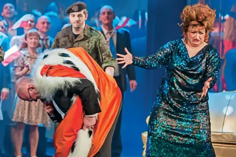 Król opera Pendereckiego w pomarańczowym płaszczu z futrem kłania się nisko. Obok kobieta w błyszczącej sukni.