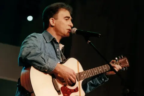 Seweryn Krajewski na scenie, gra na gitarze i śpiewa do mikrofonu. Jego muzyka wciąż porusza serca, a pytanie "czy Seweryn Krajewski żyje" jest niepotrzebne, bo jego twórczość jest wieczna.
