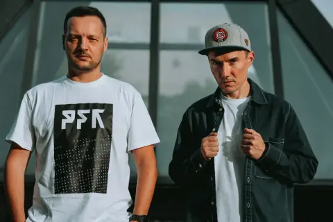 Dwóch członków Paktofoniki, jeden w białej koszulce z logo, drugi w czarnej kurtce i czapce z daszkiem.